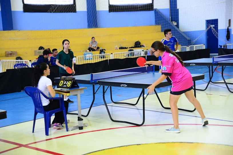 Foto 2_ Duas adolescentes jogam tênis de mesa em quadra coberta. Uma outra adolescente contabiliza o jogo sentada