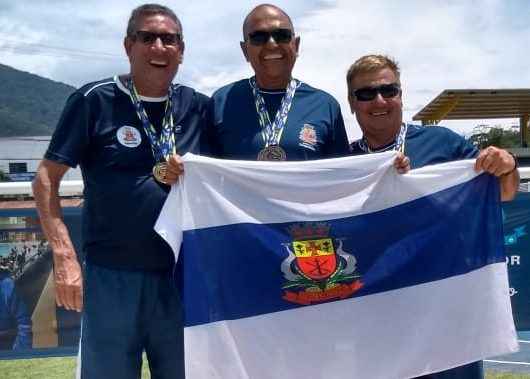 Três homens seguram bandeira de Caraguatatuba em cima do pódios nos segundo primeiro e terceiro lugar respectivamente
