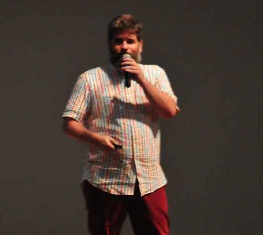 Edgar está no palco do Teatro Mario Covas. Ele veste calça vermelha de brim, blusa de tecido bege de manga curta e sapatos pretos.