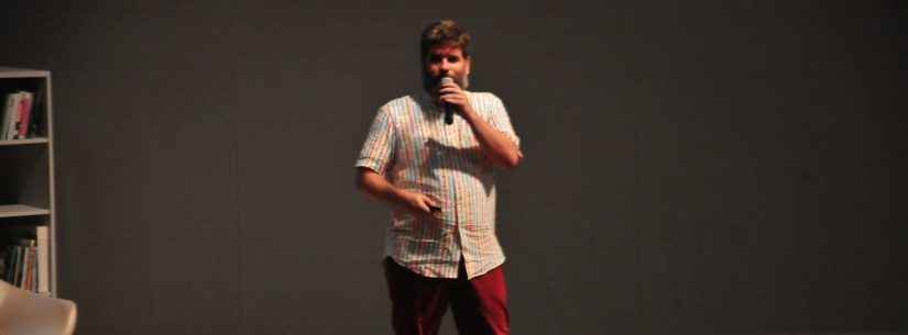 Edgar está no palco do Teatro Mario Covas. Ele veste calça vermelha de brim, blusa de tecido bege de manga curta e sapatos pretos.