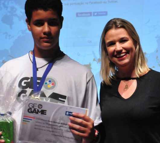 De pé, no palco do Teatro Mario Covas, da esquerda para a direita, a supervisora Karina Soares, o vencedor, Kauan, que segura o prêmio com a mão direita e o certificado com a mão esquerda com a medalha no pescoço, a secretária Márcia Paiva e a coordenadora do Apoio Pedagógico, Alessandra Ciaca.