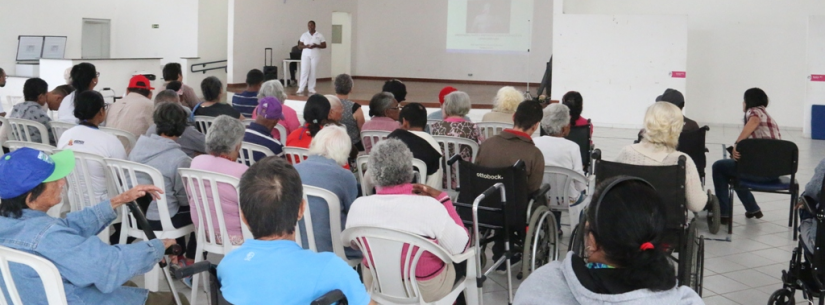 Várias pessoas sentadas no salão do Ciapi, assistindo palestra, enquanto palestrante está em pé, em cima do palco, está vestida de calça e camiseta branca.