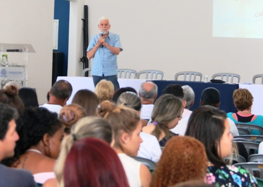 Pessoas sentadas assistindo palestra do advogado, que está em pé em cima do palco com microfone. Ao seu lado, sentada na mesa, está a promotora