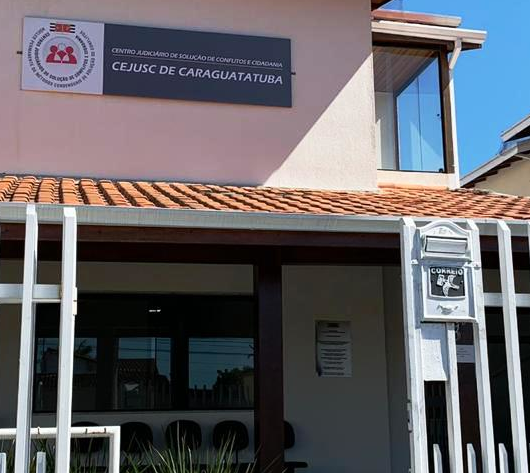 Fachada do Cejusc, sobrado da cor rosê, com portão branco de alumínio e gramado na varanda. Em cima, placa escrito Cejusc de branco e fundo azul