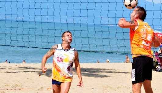 Dupla de Futevôlei vestidas com uniforme na praia disputando uma competição de um campeonato anterior. Um jogador da dupla lança a bola para a dupla adversária