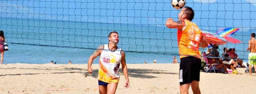 Dupla de Futevôlei vestidas com uniforme na praia disputando uma competição de um campeonato anterior. Um jogador da dupla lança a bola para a dupla adversária